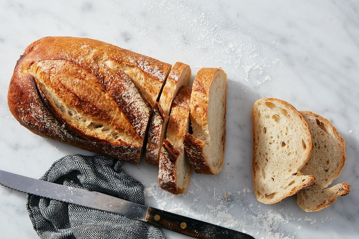 A sliced rustic sourdough loaf on a marble surface, open crumb and dark crust, with a bread knife and cloth