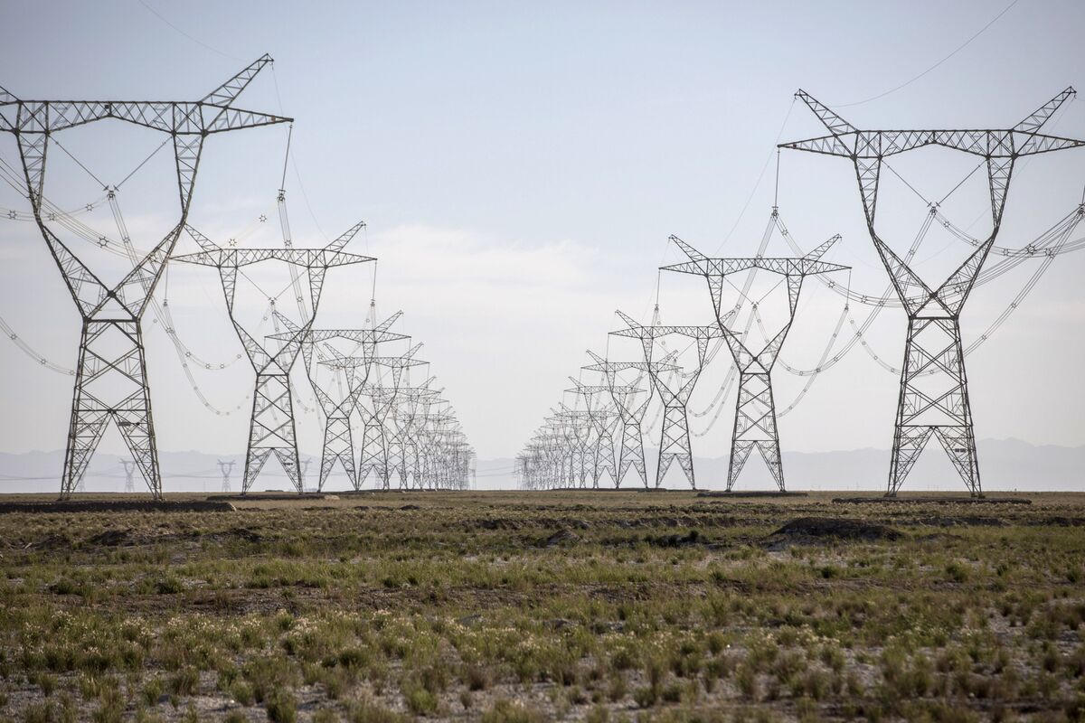 Rows of ultra-high-voltage transmission towers marching across flat scrubland toward distant hills in western China under a pale sky.