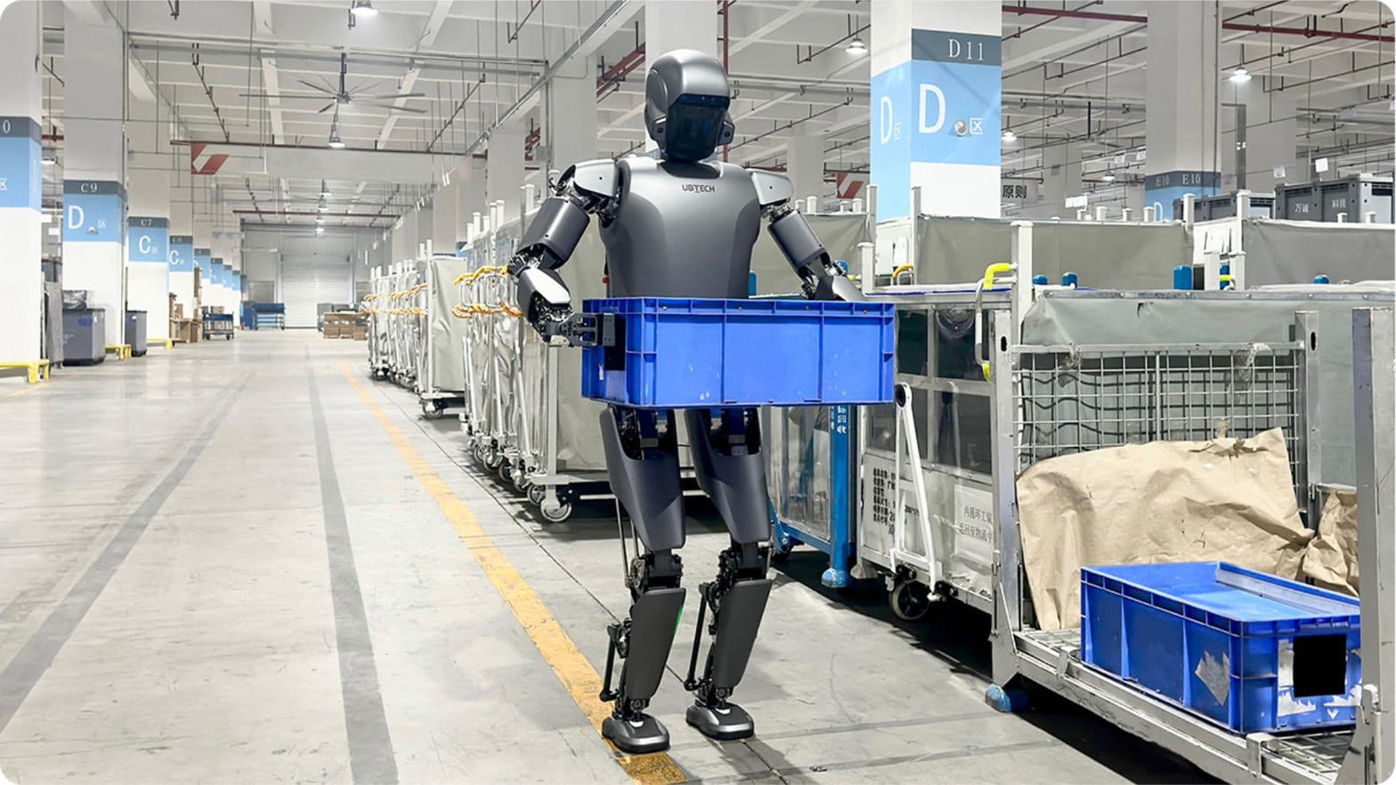 A UBTECH Walker humanoid robot carrying a blue plastic crate along a marked aisle on a Chinese factory floor, with material trolleys lining the bay behind it.