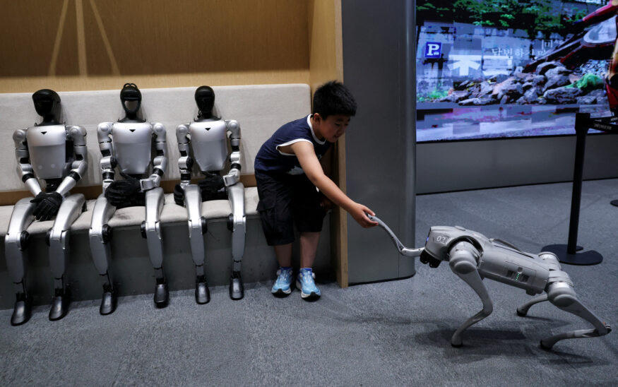 Three Unitree humanoid robots seated on a bench at a public exhibition while a young boy leans around a pillar to touch a Unitree quadruped robot walking toward him.